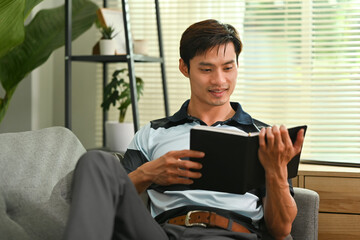 Carefree millennial man relaxing on comfortable couch and reading book, spending quality time at home