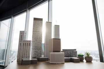 Fototapeta premium Architectural bureau office with panoramic windows and blurry cityscape. Building complex prototype project of residential or business district, city model. 3D city buildings on a wooden table.