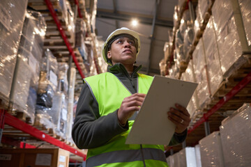 Low angle portrait of black woman wearing hardhat in storage warehouse and holding clipboard