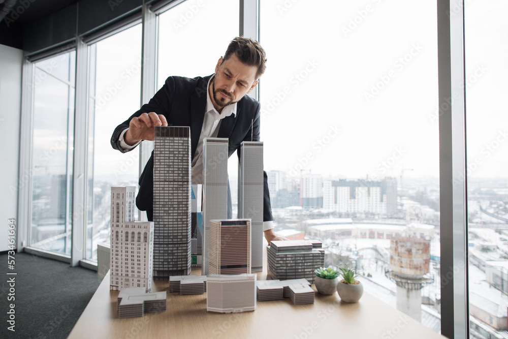 Wall mural Confident businessman real estate agents in black stylish formal suit standing while touching 3d model skyscraper of city architecture design at office with panoramic city view.