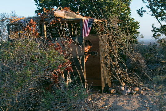 A little boy plays in a jungle inside a deserted soldier outpost, he takes aim like a soldier with a gun like stick, a USA national flag flies from the top of the outpost.