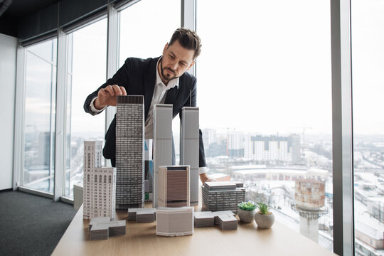 Confident businessman real estate agents in black stylish formal suit standing while touching 3d model skyscraper of city architecture design at office with panoramic city view. - Powered by Adobe