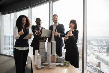 Team of diverse four multiethnic businesspeople having meeting in boardroom at office with panoramic windows blurry cityscape standing near table with skyscraper building maquette showing thumbs up