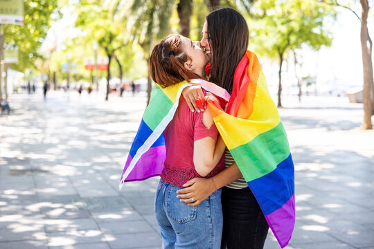 Lesbian couple covered in rainbow colored flag hugging each other on footpath