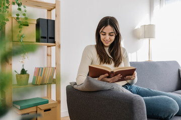 Smiling woman reading book sitting on sofa in living room
