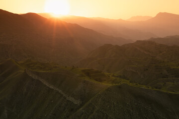 Majestic landscape - silhouette of high mountain green ridges in bright golden sunlight with sunbeams and pastel mist in warm sunset of summer day, panorama view. Joyful trekking in Caucasus mountain.