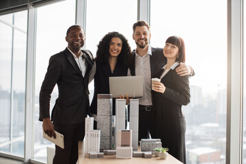 Happy smiling successful diverse multiracial business coworkers, posing on camera during meeting in office room working with city buildings model. Bright people work as urban planners.
