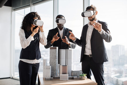 Multiracial business people, using VR glasses, making team training, gesturing, and enjoying work together, standing near table with city buildings model at office interior with panoramic city view