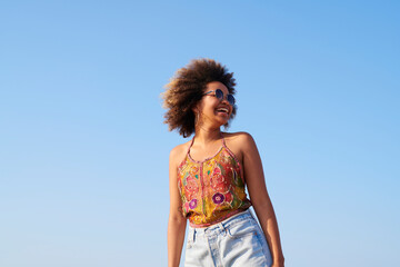 Laughing woman in sunglasses in the wind blow. Portrait of pretty cheerful woman with frizzy hair in sunglasses standing in wind blowing her hair against clear blue sky on sunny day.