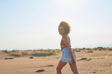 Cheerful woman in sunglasses walking in desert. Side view of happy woman with frizzy hair walking in sunny desert against blue clear sky. She is looking at camera.