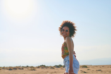 Happy woman with afro in desert. Side view of cheerful woman with afro in sunglasses, top and denim shorts laughing on sunny beach.