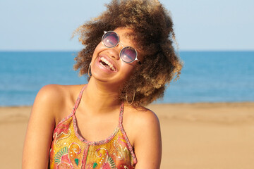 Portrait of cheerful candid girl with frizzy hair in fashionable round sunglasses and brackets wearing summer dress with bare shoulders. Unfocused seascape in background.