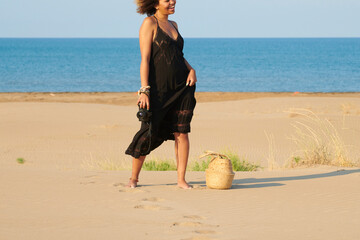 Cropped portrait of jovial dancing woman in long summer dress with professional camera with woven bag on the sand. Seascape in background.