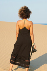 Anonymous trendy woman with camera on beach. Back view of ethnic woman in stylish black dress standing on beach with camera in sunlight