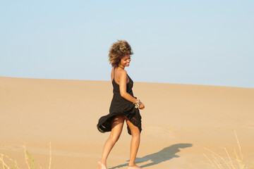 Playful black woman in dress on beach. Dancing happy woman with Afro hairstyle wearing black dress and having fun on sandy beach on blue sky background