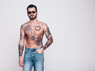 Fototapeta premium Portrait of handsome confident stylish hipster lambersexual model in jeans. Sexy modern man. Naked torso with tattoos. Fashion male posing in studio. Isolated on white. In sunglasses