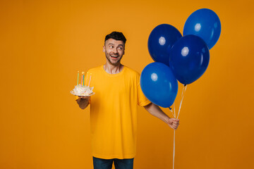 Excited man holding birthday cake and blue air balloons isolated over yellow wall