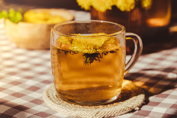 Dandelion flower healthy tea in glass teapot and glass cup on table. Delicious herbal Hot tea from fresh dandelion flowers at home at summer day. Green clearing. Bouquet of dandelions petals