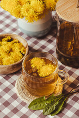 Dandelion flower healthy tea in glass teapot and glass cup on table. Delicious herbal Hot tea from fresh dandelion flowers at home at summer day. Green clearing. Bouquet of dandelions petals