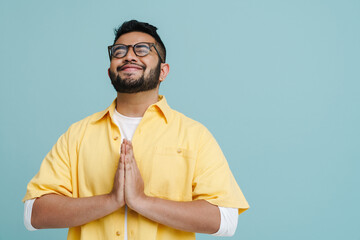 Indian man making pray gesture while posing with eyes closed isolated over blue background