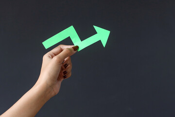 Hand hold arrow cutted from solid sheet of green paper and curved up of one side showing growth of stock market or up direction isolated on dark background with copy space. 