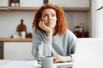 Charming ginger-headed female with glasses on head looking up with thoughtful facial expression, sitting at kitchen table with coffee and laptop, writing study plan and creating schedule for students