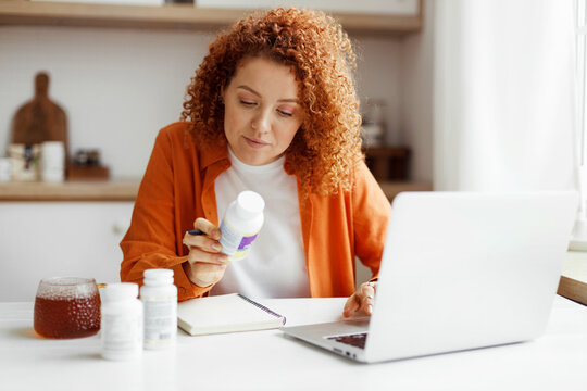 Redhead nutritionist working from home on laptop at her article about deficiency of food supplements and its influence on human body and health sitting at kitchen table with vitamin bottle