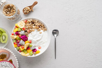 Heathy tropical fruit yogurt bowl with granola, mango, kiwi, bananas, dragon fruit and pomegranate on light grey background, top view, copy space