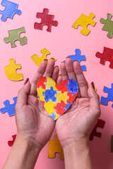 Hands hold colorful puzzle heart shape isolated on colorful puzzle on pink background. World Autism...