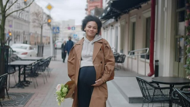 Pregnant young african american woman walks through the street in a happy mood with flowers in her hands