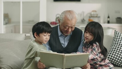 asian grandpa and two grandchildren sitting on couch at home reading a book together - Powered by Adobe