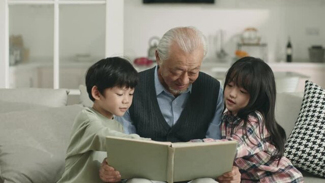 asian grandpa and two grandchildren sitting on couch at home reading a book together