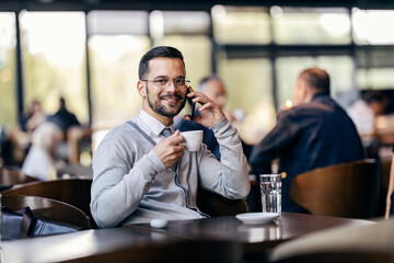 A young happy man in smart casual is sitting in cafe and drinking coffee while having a phone call.
