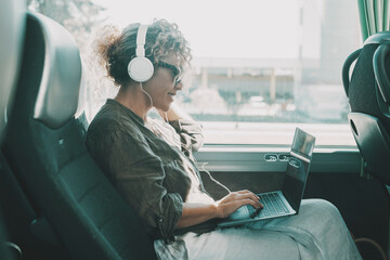 Young woman work on laptop with headset sitting on bus seat. Travel business lifestyle female people. Modern lady smiling and enjoying trip as passenger on transport public vehicle. Technology online - obrazy, fototapety, plakaty