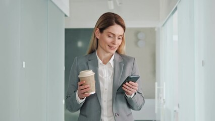 Closeup happy businesswoman using mobile phone at office hall holding morning coffee. Smiling woman browsing internet on smartphone in office. Portrait of business woman reading messages on cellphone.