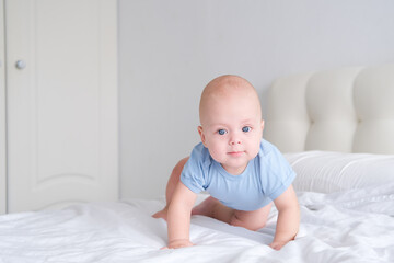 smiling boy with big blue eyes in a bodysuit learns to crawl on white bedding. Healthy newborn baby