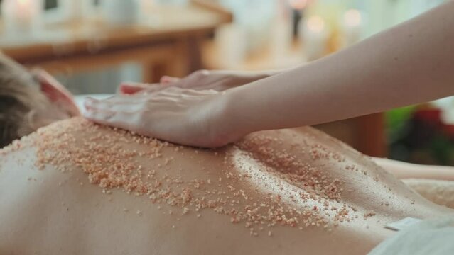 Unrecognizable massage therapist rubbing salt scrub on back of young woman relaxing during spa procedure