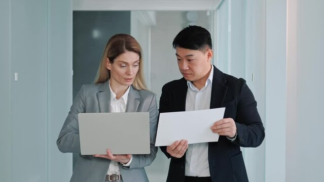 Multicultural business partners working together looking at laptop screen. Two business people working in office hallway reading and discuss asian investment project, analyze the stock market.