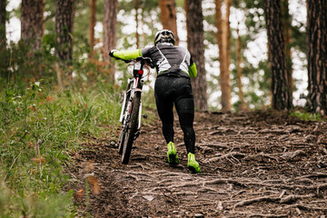 back cyclist walking uphill with mountain bike. forest trail with tree roots