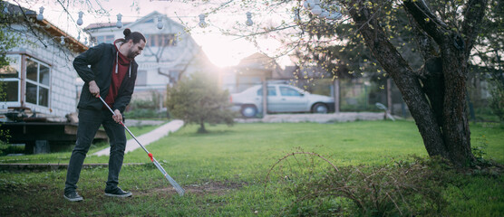 A young man works in the garden, loosens the ground with a fan rake, removes dry grass from the lawn