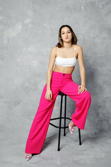 Slender young woman in wide cherry pink trousers and white tube top sitting on tall chair