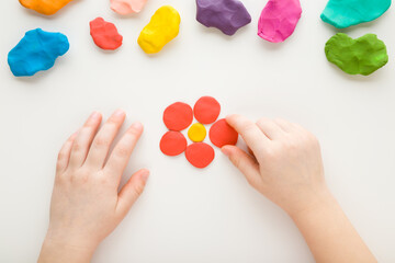 Baby girl hands modeling red flower head from clay on white table background. Point of view shot. Closeup. Toddler development. Top down view.