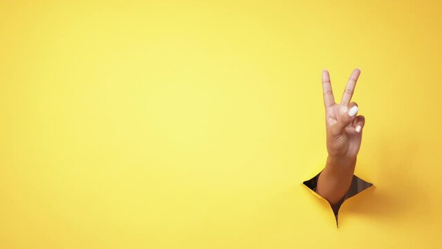 Peace gesture. Victory signal. Winner support. Female hand showing V sign in yellow breakthrough hole ripped paper wall advertising copy space background.