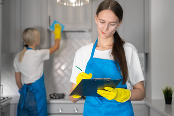 Young woman from a professional cleaning company in uniform notes and writes down a list of the work done on cleaning the room in a tablet. Stove is being laid in the background.