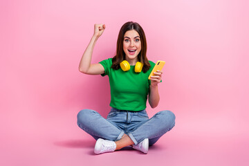 Full size portrait of delighted funky girl sit floor hold smart phone raise fist success isolated on pink color background