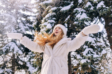 Young woman dressed in a light fur coat plays with snow in winter