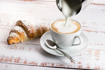 Making cappuccino with milk frother on wooden table with croissant.