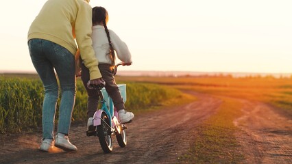 mother teaches child ride two-wheeled bicycle sunset. fun weekend by bike. child's dream kid. child...