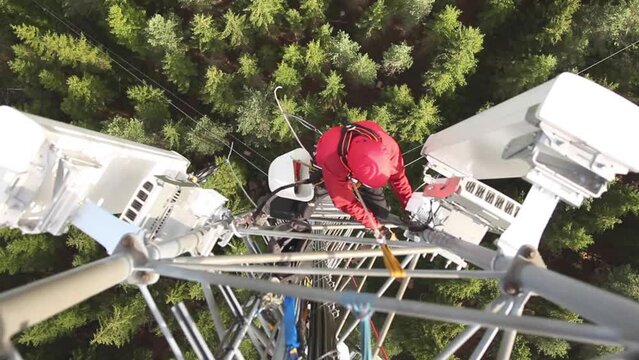 A high-angle view of a telecommunications technician working at height on a telecom tower, performing maintenance on 5G antenna.