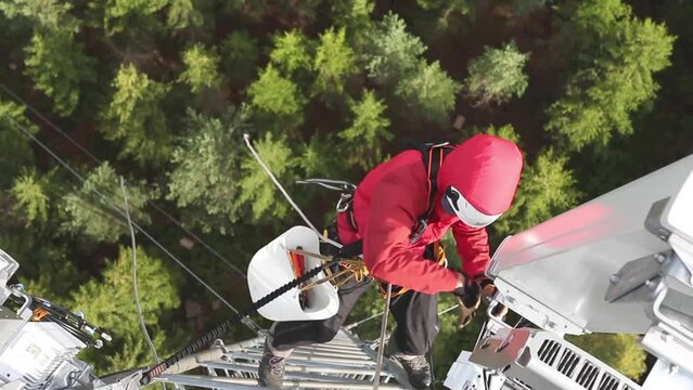 A high-angle view of a telecommunications technician working at height on a telecom tower, performing maintenance on 5G antenna.